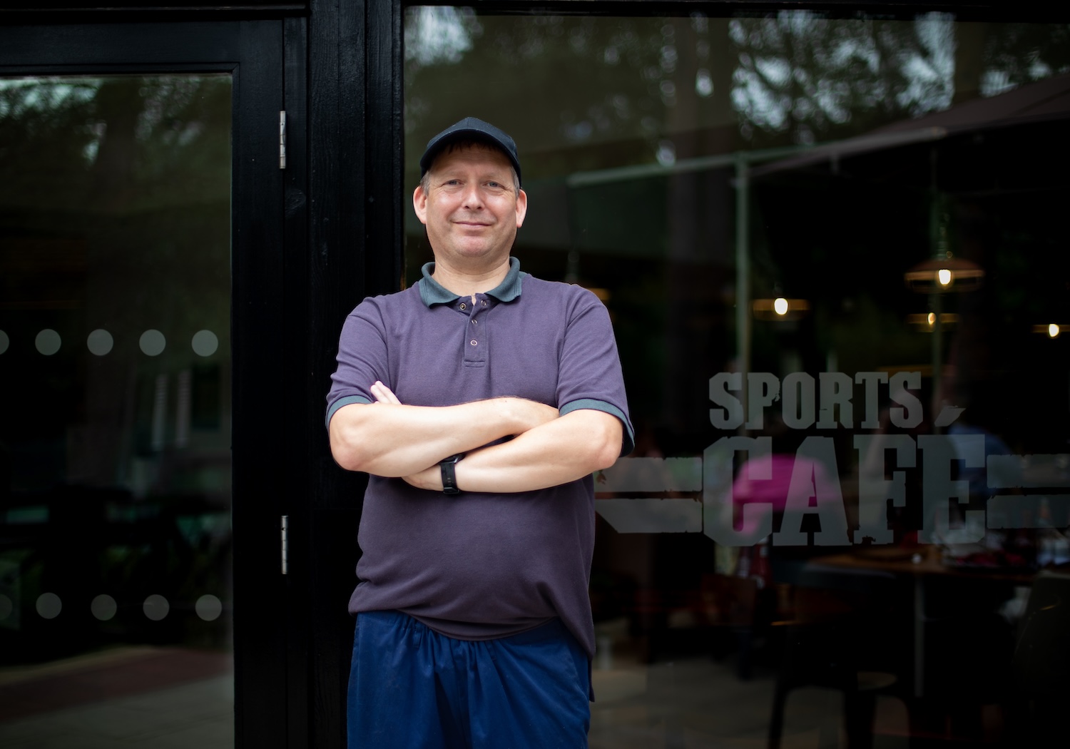 Man in front of Sports Cafe, arms crossed, wearing a cap and polo shirt.