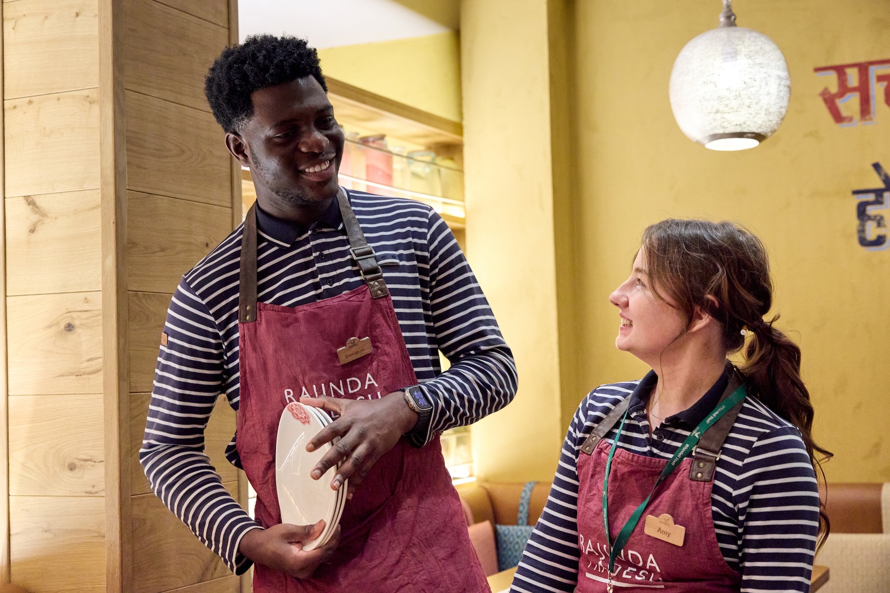 Two restaurant employees smiling, wearing aprons and striped shirts.