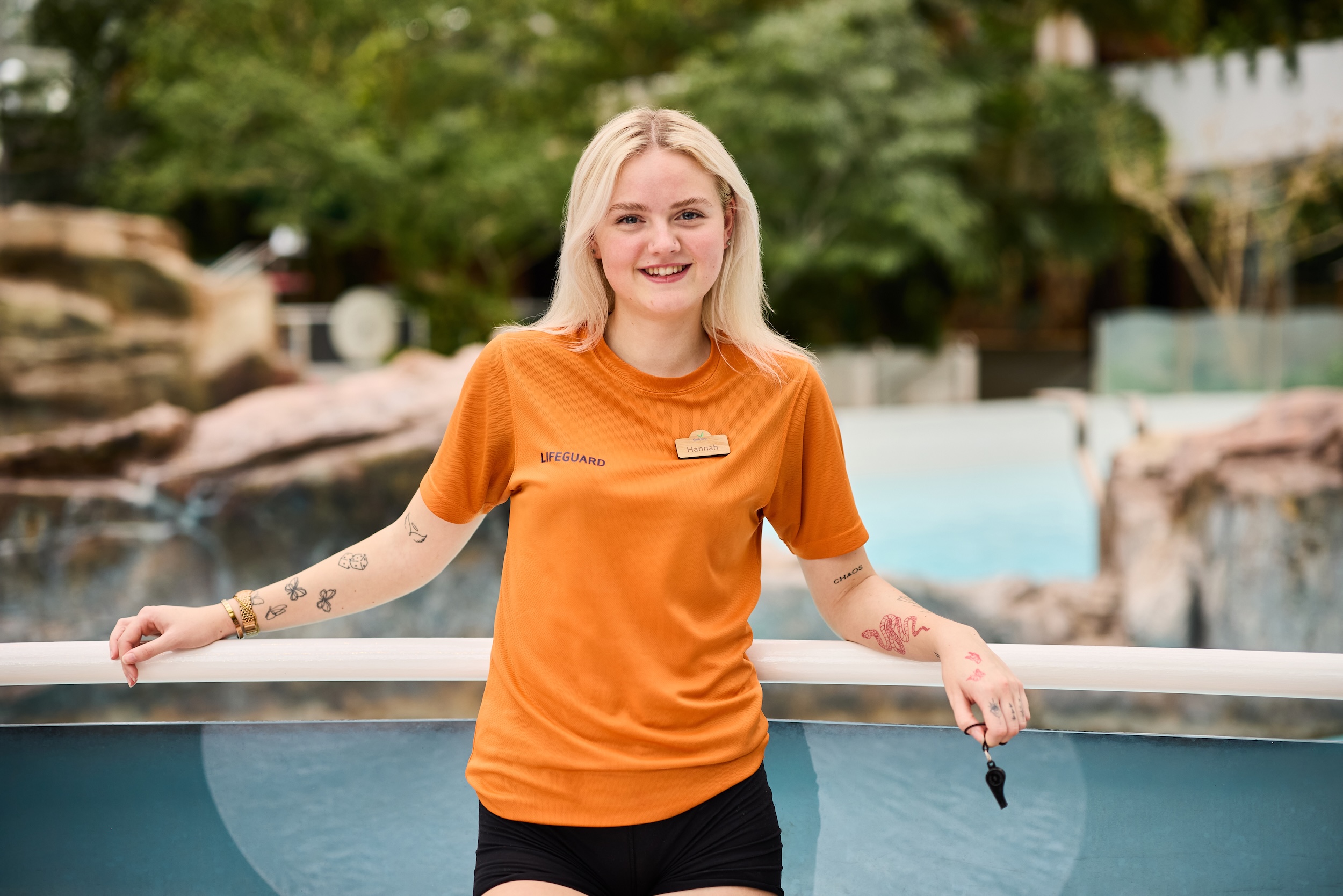 Blonde lifeguard in orange shirt, tattoos, and whistle.