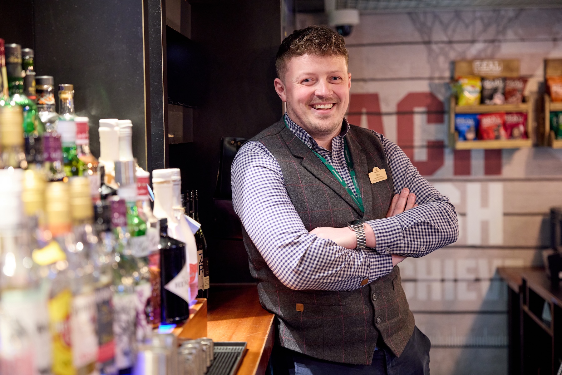 Man smiling at the bar, arms crossed, wearing a vest and name tag.
