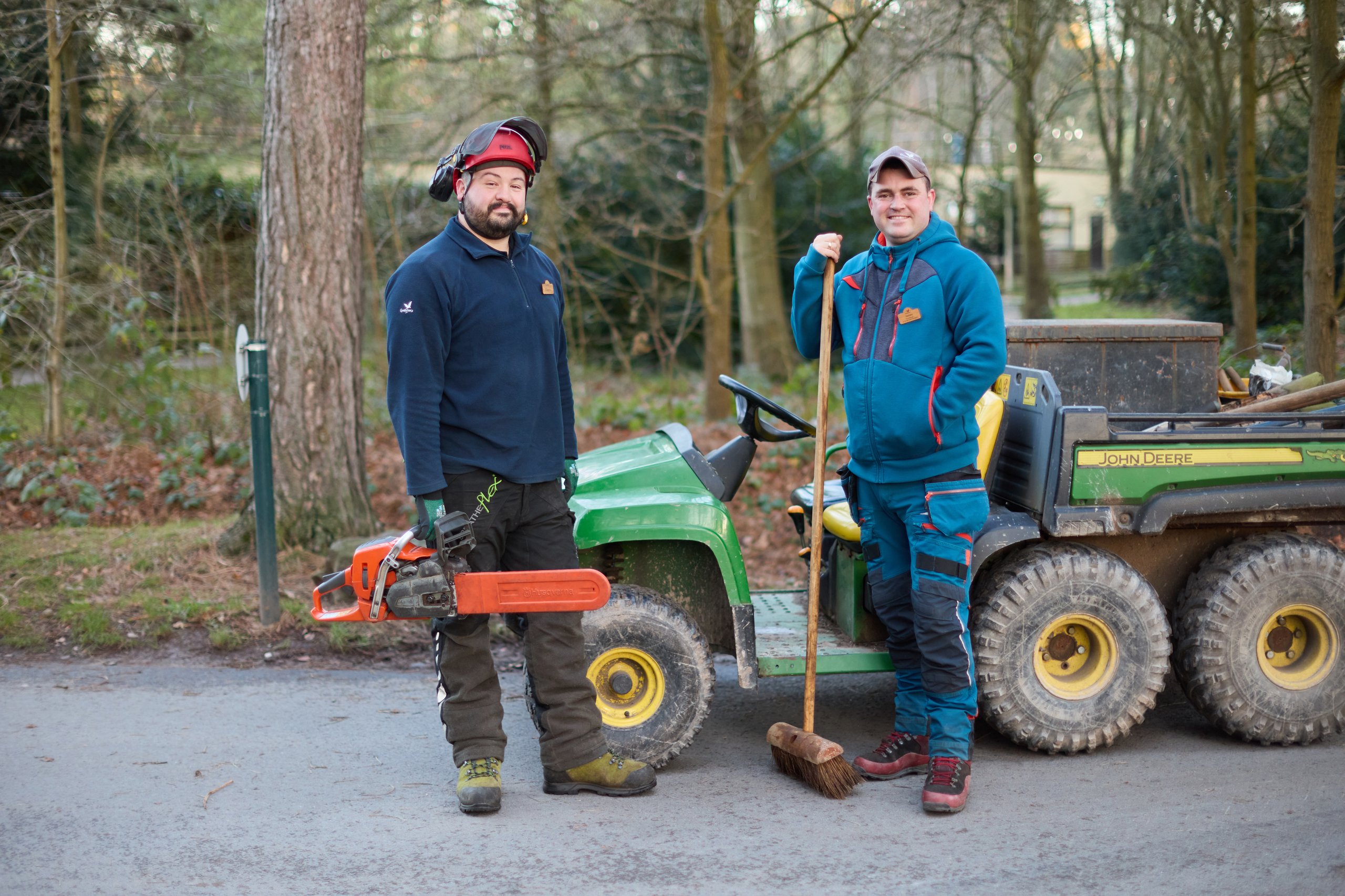 Two men in work attire standing near a John Deere vehicle, one with a chainsaw, the other with a broom.