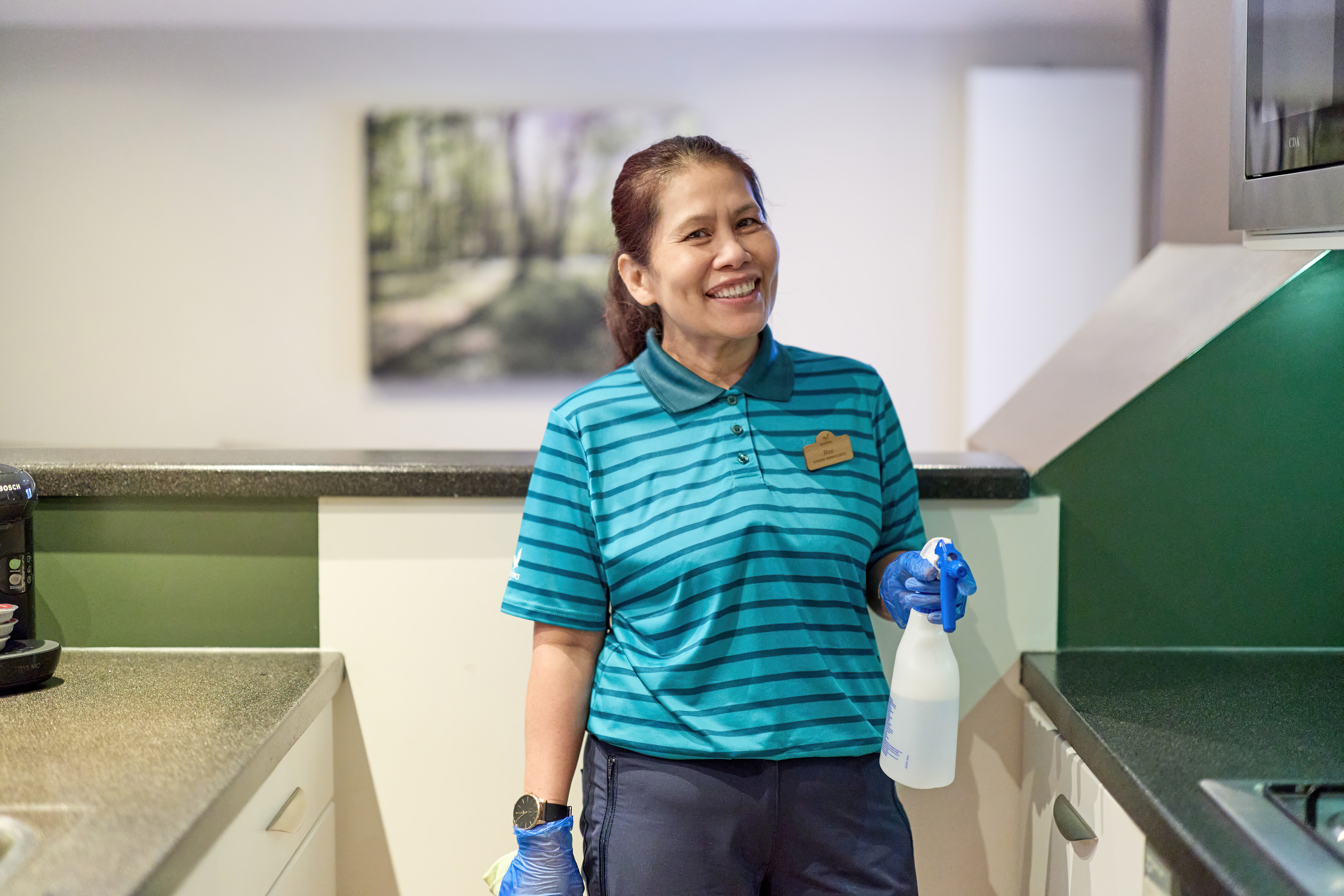 Housekeeper in uniform holding spray bottle, smiling.