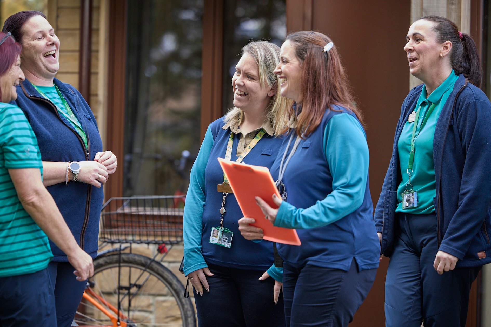 Four housekeepers in matching uniforms laugh and talk outside a building, one holding an orange clipboard.