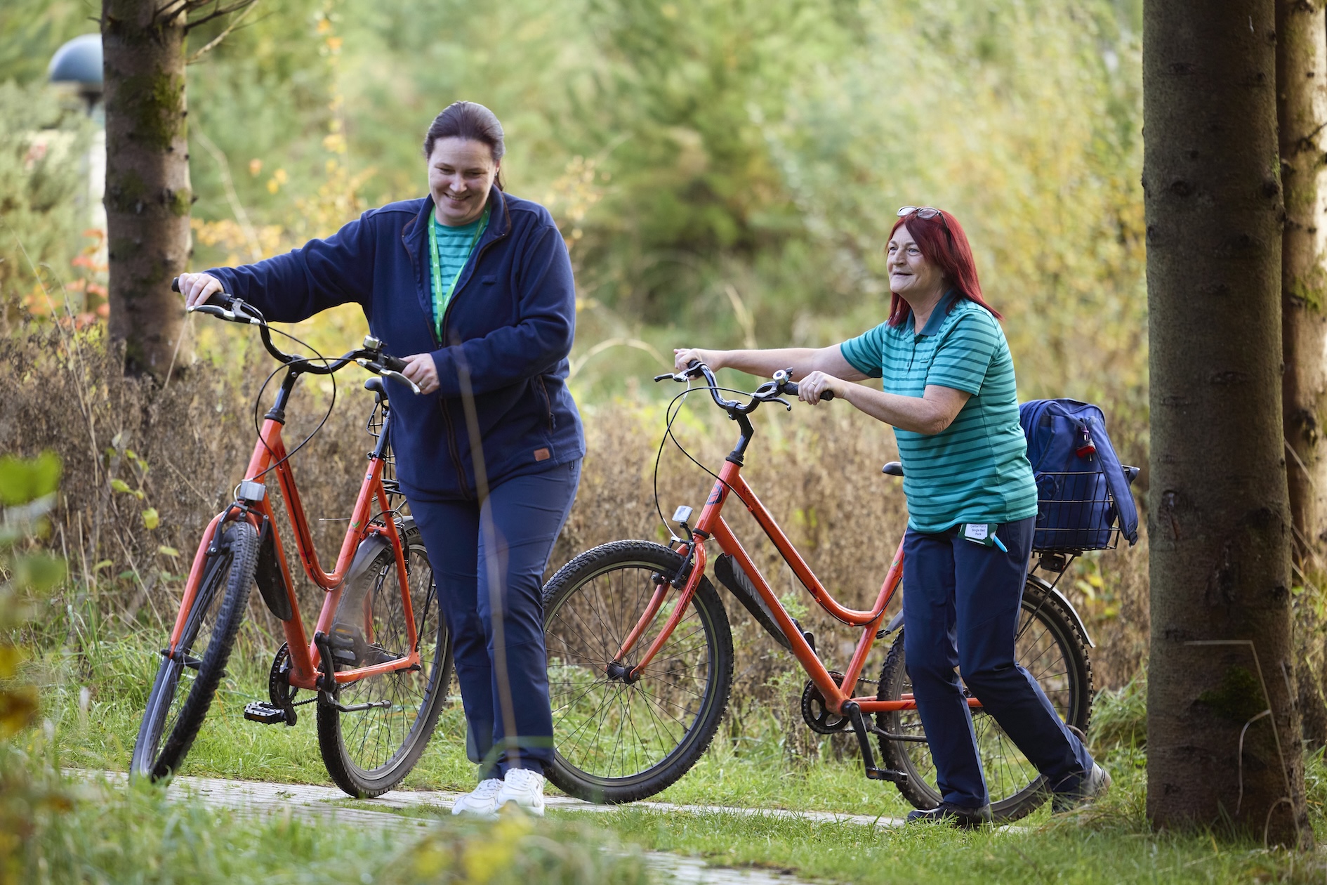 Two women with bicycles walk on a path surrounded by trees and foliage. One woman smiles while looking at the other, who is also smiling and looking ahead.