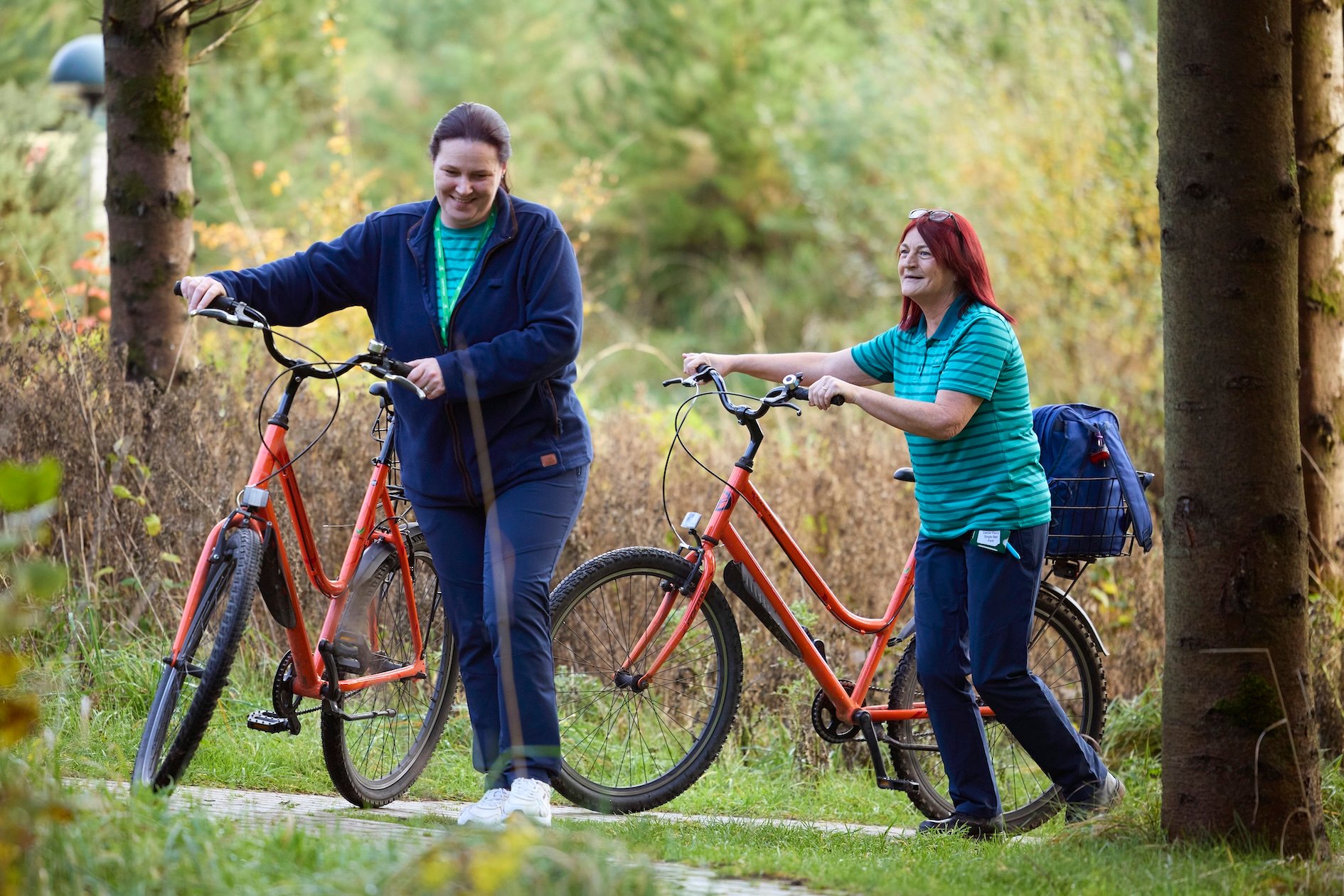 Two women with bicycles walk on a path surrounded by trees and foliage. One woman smiles while looking at the other, who is also smiling and looking ahead.