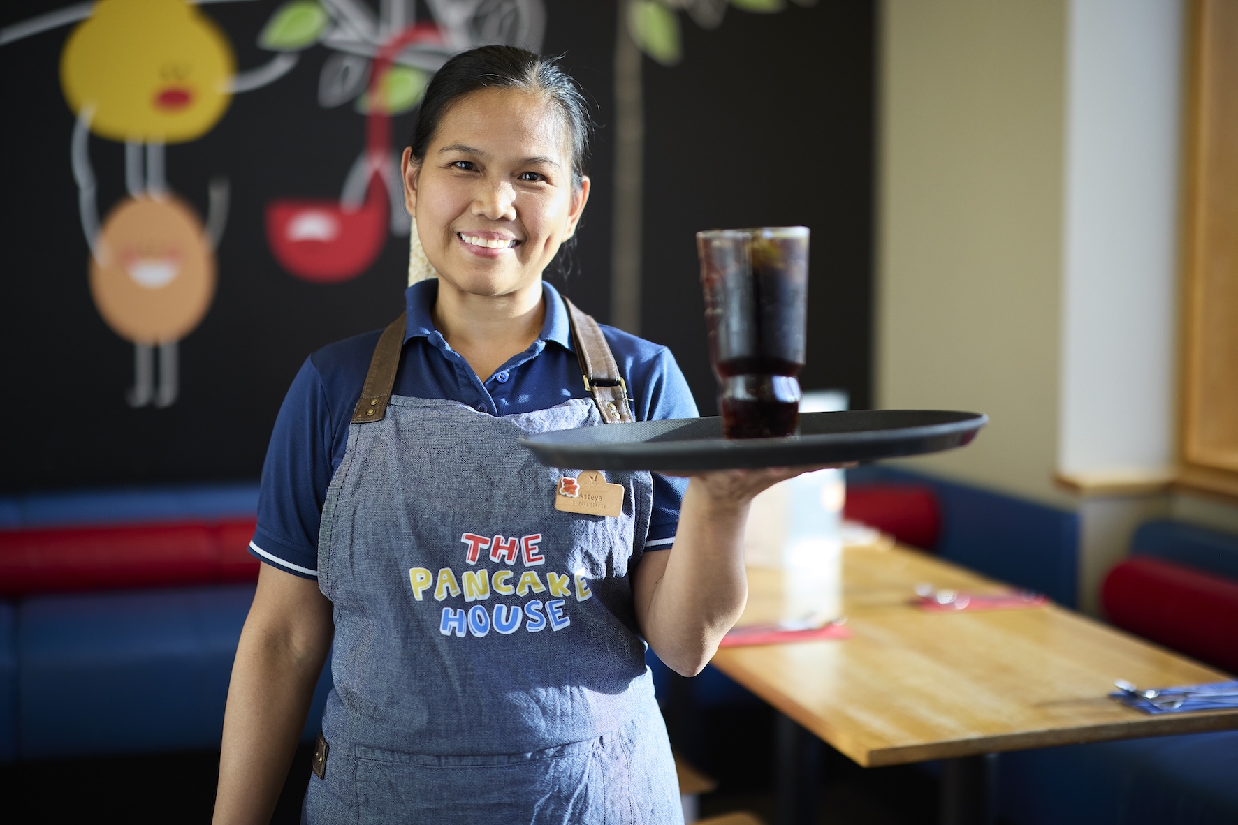 A server at The Pancake House smiles while holding a tray with a glass of dark soda and ice.